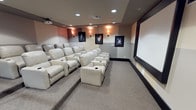 This private home theater is thoughtfully designed to deliver a true cinematic experience. The room is arranged with tiered rows of plush, cream-colored leather reclining seats, each equipped with built-in cup holders for added convenience. The tiered layout ensures unobstructed sightlines to the large projection screen that dominates the front wall, framed with sleek border trim for a polished, theater-quality finish. Recessed ceiling lighting and warm wall sconces create a soft, ambient glow that sets the mood without distracting from the screen. The walls are adorned with framed black-and-white portraits of classic Hollywood icons, lending the space a nostalgic, cinematic charm. Neutral-toned carpeting runs throughout, absorbing sound and contributing to the room's excellent acoustics. The combination of neutral gray and warm accent wall colors adds depth and sophistication to the interior. A ceiling-mounted projector completes the setup, ensuring a high-quality viewing experience. Every detail of this space has been curated to blend comfort, style, and function into a premier private screening room.