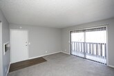 This single-story residence offers a practical and well-organized layout across several distinct areas. The living room serves as the central gathering space, featuring plush wall-to-wall carpeting, neutral gray walls, and a large sliding glass door fitted with vertical blinds that allows natural light to fill the room. An open pass-through connects the living area to the kitchen, which is equipped with white cabinetry, a full-size refrigerator, an electric range, and durable wood-look flooring that contrasts with the carpeted areas. A ceiling fan with integrated lighting is present in the kitchen as well.

The unit includes two bedrooms, both carpeted and painted in soft, neutral tones with textured ceilings and ceiling fans. One bedroom features a built-in closet with shelving, while the other offers access to additional storage. Each room provides a comfortable and functional space. The bathroom is finished with wood-look flooring, a white vanity with updated hardware, and a tub-shower combination. Throughout the unit, consistent gray paint tones and white trim create a clean, cohesive aesthetic.