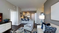 This well-appointed single-floor residence showcases a thoughtfully designed open-concept layout that seamlessly connects the living and kitchen areas. The living space is anchored by rich, dark hardwood flooring that contrasts elegantly with warm neutral-toned walls, creating a refined and inviting atmosphere. A plush area rug with a bold botanical pattern in blue and white grounds the seating area, which features a patterned accent chair and a generously sized sofa dressed with navy blue throw pillows — together establishing a cohesive and sophisticated color scheme throughout.

The kitchen is visible from the living area and offers a clean, contemporary aesthetic with white cabinetry, stainless steel appliances, and pendant lighting that illuminates the space with a warm glow. A white breakfast bar with stylish navy blue bar stools serves as both a functional dining perch and a natural visual divider between the kitchen and living areas. Framed artwork on the walls adds a curated, gallery-like quality to the space. The overall ambiance is modern and polished, with careful attention to detail in the furnishings, lighting, and decor — making this a comfortable and visually striking living environment.