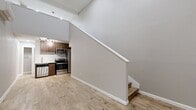 This two-story, 1-bedroom, 1-bathroom residence offers a smart and efficient loft-style layout spread across two levels. The lower floor serves as the main living area, immediately impressing with soaring ceilings that lend a sense of openness far beyond the unit's footprint. Light wood-look flooring runs throughout, complementing the neutral gray walls and clean white trim. A compact yet well-appointed kitchen anchors one end of the lower level, featuring warm walnut-toned cabinetry, a white subway tile backsplash, granite-style countertops, and a full suite of stainless steel appliances including a refrigerator, range, and microwave. A sleek black-framed glass entry door adds a contemporary touch to the main level. The staircase, finished with a crisp white railing, leads to the upper floor, which houses the bedroom. This private retreat is outfitted with a wall-mounted mini-split HVAC unit, a horizontal window for natural light, and recessed lighting. The bathroom features wood-look tile flooring, a walnut vanity with a stone-topped vessel sink, and a full soaking tub surrounded by elegant marble-style tile surround, creating a refined and spa-like atmosphere.