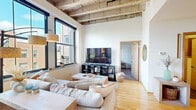 This single-floor urban loft showcases a compelling blend of industrial heritage and contemporary living. The defining architectural feature is the striking exposed concrete beam ceiling in the main living area, which lends the space a raw, warehouse-inspired character while soaring to impressive heights. Large, black-framed windows flood the interior with abundant natural light and frame sweeping city views, creating a bright and energizing atmosphere throughout.

The open living area is anchored by warm hardwood flooring that flows seamlessly through the main spaces. A generously sized seating arrangement makes the living room an inviting hub for relaxation and entertainment, complemented by a dedicated media setup and additional shelving for storage and display.

The kitchen is well-appointed with light maple cabinetry, dark granite countertops, stainless steel appliances including a refrigerator, range, and built-in microwave, and a tile backsplash. A substantial island provides additional prep space and casual seating.

A versatile secondary room with a tall city-facing window serves as a functional home office, complete with ample natural light ideal for focused work. The bedroom offers a calm, carpeted retreat with a window overlooking the urban surroundings, providing a quiet contrast to the loft's open, airy communal spaces.