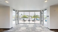 Located at 12850 SW Ash Ave in Tigard, Oregon, this single-floor community space offers a thoughtfully designed environment that seamlessly blends indoor comfort with outdoor living. The interior living area features patterned carpet flooring in neutral grey tones, clean white walls with warm yellow accent walls, and large windows that draw in generous amounts of natural light. Glass doors open directly onto an outdoor terrace, creating a smooth transition between the two areas. The outdoor terrace is a standout feature of the space, finished with large-format light stone tile and framed by a dark-paneled accent wall that adds a bold, contemporary contrast. Comfortable seating — including a woven sofa, teal-cushioned lounge chairs, and sculptural side tables — creates an inviting atmosphere for gathering. Planters with flowering plants and a striking mature tree add organic warmth to the setting. A partial skylight structure overhead allows natural light to filter into the terrace while providing shelter. The overall ambiance is modern and refined, balancing clean architectural lines with carefully curated décor, making it a welcoming communal retreat in the heart of Tigard.
