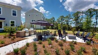 Located on Johns Island, SC, this outdoor community amenity space offers a well-appointed environment designed for relaxation and social gathering. The grounds are thoughtfully landscaped with native plantings, pine straw mulch beds, and flowering perennials that add color and texture throughout the property.

At the heart of the outdoor area is a spacious patio featuring a built-in grilling station equipped with multiple grills, ideal for outdoor cooking and entertaining. Umbrella-shaded dining tables and Adirondack-style chairs provide comfortable seating, while a dedicated fire pit area with lounge seating creates a welcoming space for evening gatherings.

Winding gravel pathways meander through manicured green lawns, connecting the various areas of the grounds and offering a pleasant walking experience. The open lawn areas are framed by mature pine and hardwood trees, including moss-draped Southern live oaks, providing natural shade and a quintessential Lowcountry ambiance.

A black iron fence encloses portions of the grounds, adding a classic aesthetic while defining the space. The residential buildings feature traditional Lowcountry-inspired architecture with light-colored lap siding, pitched rooflines, and multi-pane windows. The overall setting benefits from abundant natural light and sweeping views of the surrounding greenery, creating a serene and inviting outdoor community environment.