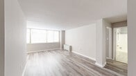 This single-floor, 2-bedroom, 1-bathroom unit offers a clean, neutral aesthetic throughout. The living area is the most expansive space in the unit, bathed in natural light from a large multi-pane window. Light gray hardwood-style flooring runs throughout the main living spaces, complementing the crisp white walls and baseboards for a bright, cohesive look. A baseboard heating unit is positioned beneath the window, ensuring warmth during cooler months.

The spacious primary bedroom features the same flooring and large windows with blinds, providing ample natural light and a comfortable, airy feel. The second bedroom is more compact but maintains the same neutral tones and consistent flooring.

The galley-style kitchen is efficiently designed with warm natural wood cabinetry lining both walls, granite-look countertops, a stainless steel sink, and white appliances including a refrigerator, range, and dishwasher. Light tile flooring distinguishes the kitchen from the rest of the unit.

The bathroom is finished with clean white subway-tile walls surrounding a full tub-shower combination, complemented by a white vanity with a solid countertop. The unit also includes two closets, offering practical built-in storage. Overall, the space presents a functional and well-maintained interior with a simple, modern palette.