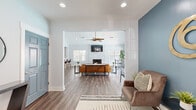 This single-floor space offers an open-concept layout that seamlessly connects the entrance, living room, dining area, and kitchen into one cohesive, functional environment. The entrance welcomes visitors with warm hardwood-style flooring, soft blue-gray walls, decorative greenery, and a full-length mirror that adds depth to the space. A distinctive gold wall accent and branded signage give the entry a polished, intentional feel.

The living area is a standout, featuring soaring vaulted ceilings, recessed lighting, and a modern ceiling fan. A stacked-stone fireplace serves as the focal point, framed by a wall-mounted television and flanked by windows that flood the room with natural light. Comfortable leather seating and a geometric area rug contribute to the room's inviting, contemporary ambiance.

The dining area sits adjacent to the living space, benefiting from the same abundant natural light and direct access to an outdoor pool area through large sliding glass doors — offering pleasant views and a strong indoor-outdoor connection.

The kitchen is equipped with dark cabinetry, stainless steel appliances, and a granite-topped breakfast bar with bar seating, making it both practical and stylish. Throughout the space, consistent hardwood-style flooring, neutral tones, and thoughtful décor create a modern, cohesive atmosphere.