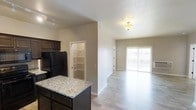 This single-floor residence showcases an open-concept layout that seamlessly connects the kitchen and living areas. The kitchen is outfitted with dark espresso cabinetry, granite countertops on a central island, and black appliances, including a full-size refrigerator. A walk-in pantry with built-in shelving is conveniently accessible directly from the kitchen, offering generous storage space.

Light wood-tone laminate flooring runs throughout, providing a clean, cohesive look that enhances the sense of openness. Neutral warm-beige walls complement the overall aesthetic, creating a welcoming and versatile backdrop.

The living area receives abundant natural light through a sliding glass door that leads to an outdoor balcony, making the space feel airy and bright. Overhead lighting, including a flush-mount ceiling fixture and a track light in the kitchen area, ensures the space is well-lit throughout the day and evening. The open flow between the kitchen and living space makes this unit well-suited for both everyday living and entertaining.