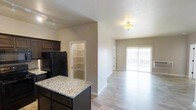 This single-floor residence showcases an open-concept layout that seamlessly connects the kitchen and living areas. The kitchen is outfitted with dark espresso cabinetry, granite countertops on a central island, and black appliances, including a full-size refrigerator. A walk-in pantry with built-in shelving is conveniently accessible directly from the kitchen, offering generous storage space.

Light wood-tone laminate flooring runs throughout, providing a clean, cohesive look that enhances the sense of openness. Neutral warm-beige walls complement the overall aesthetic, creating a welcoming and versatile backdrop.

The living area receives abundant natural light through a sliding glass door that leads to an outdoor balcony, making the space feel airy and bright. Overhead lighting, including a flush-mount ceiling fixture and a track light in the kitchen area, ensures the space is well-lit throughout the day and evening. The open flow between the kitchen and living space makes this unit well-suited for both everyday living and entertaining.