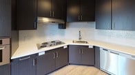 Located at 1504 Hancock St in Quincy, Massachusetts, this single-floor unit offers a thoughtfully designed layout with modern finishes throughout. The living areas are characterized by light wood-tone flooring and clean white walls, creating a bright, open atmosphere. Large windows frame expansive views of the Quincy skyline and surrounding neighborhood, flooding the space with natural light.

The kitchen is well-appointed with dark espresso cabinetry, light countertops, and a textured tile backsplash. Stainless steel appliances — including a refrigerator, dishwasher, and built-in cooktop — provide a sleek, functional workspace. The layout transitions smoothly into the main living area, which benefits from pendant and recessed lighting.

Bedroom spaces are finished with plush carpeting and feature generous windows that continue the theme of natural light and outdoor views. Sliding closet doors offer practical storage solutions.

The bathrooms are finished with wood-look tile flooring and dark vanity cabinetry topped with light countertops. One bathroom includes an accessible roll-in shower with grab bars and a handheld showerhead, reflecting a thoughtful, inclusive design approach. Chrome hardware and clean lines complete the contemporary aesthetic throughout.
