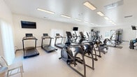 Located at 7619 Sylvan Towne Dr in Sylvania, Ohio, this fitness center offers a generously sized, single-floor workout facility designed to accommodate a wide range of exercise needs. The space features a clean, modern aesthetic with crisp white walls, light-toned flooring, and a bright ceiling lined with recessed rectangular panel lighting that ensures the entire room is evenly and thoroughly illuminated.

The cardio section is prominently featured and includes a row of treadmills, elliptical trainers, and stationary bikes, providing ample options for aerobic workouts. Multiple wall-mounted flat-screen televisions are strategically positioned throughout the space, allowing users to stay entertained during their sessions.

The strength training area is equally well-appointed, featuring a multi-station cable machine, dedicated weight benches, and a full rack of dumbbells organized by size. Additional free weights, kettlebells, and medicine balls are neatly arranged on storage racks, catering to functional fitness routines. Exercise mats are placed on the floor beneath key equipment to provide safety and comfort.

Natural light filters in through vertical-blind-covered windows, contributing to the open, airy ambiance of the room. A wall-mounted mirror enhances the sense of space and assists users with form checks. The layout is thoughtfully organized to provide clear circulation pathways between equipment zones, making this fitness center a functional and inviting environment for a comprehensive workout experience.