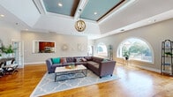 Located at 975 Westmeade Dr in Chesterfield, Missouri, this thoughtfully designed single-floor space offers a generous and open layout that balances architectural elegance with functional comfort. The main living area is immediately striking, highlighted by soaring coffered ceilings with a teal-painted inset and a statement orb chandelier that adds warmth and visual interest. Warm hardwood flooring flows seamlessly throughout the entire space, unifying each area with a cohesive, natural aesthetic.

Distinctive arched windows are a recurring architectural feature, flooding the interior with abundant natural light and offering views of the lush surrounding landscape, including a serene outdoor water feature visible from the lounge area. A floor-to-ceiling stacked stone fireplace serves as a bold focal point in the sitting area, complemented by a pair of elegant armchairs arranged for intimate conversation.

The open-concept layout accommodates a spacious main seating area, a dedicated dining space with a circular table set beneath multiple large windows, and a compact kitchen tucked in the background with rich wood cabinetry and stainless steel appliances. A quiet desk nook near a corner window provides a practical workspace. Together, these elements create a refined, welcoming atmosphere well-suited for both relaxation and gathering.