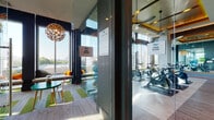This contemporary fitness facility is thoughtfully designed to offer a comprehensive wellness experience across several distinct, glass-partitioned spaces. The main gym area is lined with floor-to-ceiling windows that frame panoramic city skyline views, flooding the space with natural light throughout the day. The room is outfitted with a wide array of strength and cardio equipment, including cable machines, rowing machines, ellipticals, treadmills, and stationary bikes, all arranged on hardwood-style flooring beneath a dark, recessed ceiling accented with green and yellow architectural details and ceiling fans.

Adjacent to the main gym, the dedicated yoga and cycling studio features its own wall of expansive windows, spinning bikes arranged in rows, and an open floor area suitable for mat-based workouts. The studio's design emphasizes an airy, motivating atmosphere with natural light and city views as a backdrop.

A separate kids' room, enclosed in glass, is brightly furnished with child-sized tables, chairs, and colorful decor, providing a safe and engaging space for younger visitors. The corridor connecting these areas features bold, striped graphic wall art and terrazzo flooring accents, contributing to the facility's energetic and modern aesthetic. Together, the spaces form a well-rounded, amenity-rich fitness environment.