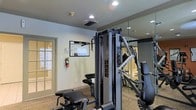 This fitness center offers a focused and functional environment designed to support a full range of exercise routines. The space is anchored by professional-grade Precor strength training equipment, including a multi-function cable machine that provides versatility for a variety of resistance exercises. Additional cardio and strength equipment is visible throughout the room, ensuring a well-rounded workout experience.

The interior features neutral-toned walls with crown molding details, giving the space a clean and polished appearance. Recessed ceiling lighting provides bright, even illumination throughout, while a large floor-to-ceiling mirror along one wall visually expands the room and allows users to monitor their form during exercise. The mirrored section also reflects the equipment, reinforcing the sense of a spacious, well-appointed facility.

Practical safety features are in place, including a wall-mounted fire extinguisher, and an equipment usage guide is posted for user reference. Ventilation is provided through ceiling-mounted air ducts, keeping the environment comfortable during workouts. A glass-paneled door offers an entry point with a view to the exterior, allowing natural light to filter into the space. The carpeted flooring adds a layer of comfort underfoot, completing this thoughtfully appointed fitness facility.