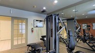 This fitness center offers a focused and functional environment designed to support a full range of exercise routines. The space is anchored by professional-grade Precor strength training equipment, including a multi-function cable machine that provides versatility for a variety of resistance exercises. Additional cardio and strength equipment is visible throughout the room, ensuring a well-rounded workout experience.

The interior features neutral-toned walls with crown molding details, giving the space a clean and polished appearance. Recessed ceiling lighting provides bright, even illumination throughout, while a large floor-to-ceiling mirror along one wall visually expands the room and allows users to monitor their form during exercise. The mirrored section also reflects the equipment, reinforcing the sense of a spacious, well-appointed facility.

Practical safety features are in place, including a wall-mounted fire extinguisher, and an equipment usage guide is posted for user reference. Ventilation is provided through ceiling-mounted air ducts, keeping the environment comfortable during workouts. A glass-paneled door offers an entry point with a view to the exterior, allowing natural light to filter into the space. The carpeted flooring adds a layer of comfort underfoot, completing this thoughtfully appointed fitness facility.