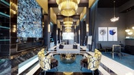 This single-floor space is a masterclass in contemporary luxury design, commanding attention with its soaring double-height ceilings and a cascading series of large, tiered gold chandeliers that bathe the entire area in warm, dramatic light. The interior palette is defined by deep charcoal and near-black walls, which are artfully contrasted by gleaming gold and metallic accents throughout.

The central lounge area features a plush, neutral-toned sofa adorned with dark accent pillows, flanked by a pair of bold gold-and-white floral-patterned armchairs. A cylindrical gold and glass cocktail table anchors the seating arrangement, which rests atop a rich teal area rug set against polished black-and-white marble flooring.

A large, vibrant abstract wall installation in deep blues and teals serves as a dramatic focal point on one side, adding an artistic dimension to the space. Tall dark drapery panels line the walls, enhancing the sense of depth and elegance. Vertical accent lighting and illuminated column details further define the corridor-like depth of the space, drawing the eye toward a softly lit reception area at the far end. The overall ambiance is bold, sophisticated, and unmistakably upscale.