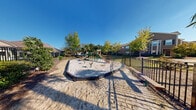 Located at 2000 Central Parkway in Montgomery, Alabama, this outdoor playground area offers a well-equipped recreational space designed for children of all ages. The play area features a generous sand-filled base that provides a safe and cushioned surface throughout. At the center of the space stands a multi-component play structure with multiple slides, climbing apparatus, and a covered canopy section, all finished in green and tan tones that blend naturally with the surrounding landscape.

Complementing the main structure are spring riders anchored in the sand, offering additional play options for younger children. The entire play area is enclosed by a sturdy black wrought-iron fence, providing a secure perimeter that enhances safety while maintaining an open, airy feel.

The playground is framed by a mix of deciduous trees and palm trees that provide partial shade and a welcoming, park-like atmosphere. A wooden boardwalk-style pathway runs along one edge of the fence, offering convenient access to the space. The surrounding landscape includes well-maintained greenery, ornamental shrubs, and views of the neighboring residential buildings. A community amenity building with a pool is visible in the background, suggesting this playground is part of a broader residential community. Under clear blue skies, the space presents a bright and inviting environment for outdoor recreation.