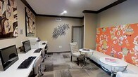 This thoughtfully designed single-floor space serves as a functional yet visually engaging business center or leasing office. The room features a workstation area along one wall, equipped with computer terminals and task chairs, making it well-suited for residents or guests who need a dedicated space for work or administrative tasks.

At the heart of the room, a round white accent table is paired with a tufted white accent chair, creating an inviting spot for casual meetings or quiet reading. A sleek white bench alongside the table adds additional seating and reinforces the room's clean, contemporary aesthetic.

The décor is a notable highlight of the space. Bold, colorful artwork adorns the walls, including a vibrant orange-toned canvas print that serves as a striking focal point. A sculptural metal wall piece with a starburst-like design adds an artistic, dimensional element to the neutral-toned walls. Crown molding with dark trim accents frames the room elegantly, adding a refined architectural detail.

The flooring consists of a patterned modular carpet in a blend of grey and neutral tones, contributing texture and visual interest underfoot. Recessed lighting provides bright, even illumination throughout. A door with a slatted blind offers access to an adjacent area, completing this well-appointed and multifunctional space.