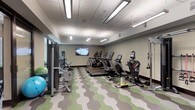 This fitness center is thoughtfully designed to support a full range of workout needs across two distinct functional zones. The main cardio and strength area is outfitted with a row of Matrix treadmills, ellipticals, stair climbers, a recumbent bike, and a multi-function cable machine, along with resistance bands and a wall-mounted flat-screen TV. The space is finished with a striking green and grey geometric patterned carpet, drop-tile ceilings with linear fluorescent lighting, and neutral-toned walls that create a clean, focused environment.

Adjacent to the main floor is a dedicated studio room featuring light hardwood-style flooring, floor-to-ceiling mirrors that amplify the sense of space, a wall-mounted TV, a full rack of dumbbells, foam rollers, and yoga mats. Large windows along one wall fill the room with natural light and offer views of an outdoor courtyard, adding an open, airy quality ideal for stretching, yoga, or strength training sessions.

The entry area of the fitness center is accented with a vibrant abstract art piece, a small beverage refrigerator, and a decorative honeycomb shelving installation that adds a modern, design-forward aesthetic. A glass-paneled door provides access to the studio area, tying the full space together in a cohesive and functional layout.