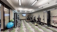 This fitness center is thoughtfully designed to support a full range of workout needs across two distinct functional zones. The main cardio and strength area is outfitted with a row of Matrix treadmills, ellipticals, stair climbers, a recumbent bike, and a multi-function cable machine, along with resistance bands and a wall-mounted flat-screen TV. The space is finished with a striking green and grey geometric patterned carpet, drop-tile ceilings with linear fluorescent lighting, and neutral-toned walls that create a clean, focused environment.

Adjacent to the main floor is a dedicated studio room featuring light hardwood-style flooring, floor-to-ceiling mirrors that amplify the sense of space, a wall-mounted TV, a full rack of dumbbells, foam rollers, and yoga mats. Large windows along one wall fill the room with natural light and offer views of an outdoor courtyard, adding an open, airy quality ideal for stretching, yoga, or strength training sessions.

The entry area of the fitness center is accented with a vibrant abstract art piece, a small beverage refrigerator, and a decorative honeycomb shelving installation that adds a modern, design-forward aesthetic. A glass-paneled door provides access to the studio area, tying the full space together in a cohesive and functional layout.
