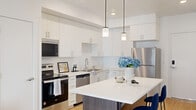 This well-appointed one-bedroom, one-bathroom residence offers a clean, modern aesthetic across its single floor. The open-concept kitchen and living area serves as the heart of the home, anchored by a striking kitchen island with a white quartz countertop, dark wood-toned base, and seating for two. Crisp white cabinetry extends to the ceiling, providing ample storage, while stainless steel appliances — including a refrigerator, range, built-in microwave, and dishwasher — lend a polished, contemporary feel. Pendant lighting above the island adds warmth and visual interest. Light wood-tone flooring flows seamlessly throughout the main living areas, enhancing the sense of openness and continuity. The living room benefits from natural light through a large window with outdoor views, creating an inviting and airy atmosphere. The spacious primary bedroom features plush carpeting, soft neutral walls, and ample room to furnish comfortably. The bathroom is finished with white cabinetry, a single-sink vanity, a large mirror, and coordinating light wood flooring. A hallway with closet access connects the spaces efficiently, completing this clean, contemporary residence designed for comfortable everyday living.