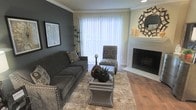 This single-story living space showcases a thoughtfully designed interior that balances comfort and contemporary style. The living room serves as the centerpiece, featuring rich hardwood flooring that adds warmth and depth to the space. A striking dark accent wall provides a bold contrast against the neutral crown molding and lighter trim, lending the room a polished, modern aesthetic.

A white-tiled fireplace anchors one wall, offering both a functional focal point and decorative appeal, accented by a decorative sunburst mirror and candle arrangements on the mantle. Natural light flows generously into the room through a large window dressed with vertical blinds, creating a bright and welcoming atmosphere.

The space is furnished with a charcoal gray sofa with nailhead trim detail, complemented by a patterned accent chair and ottoman. A plush area rug grounds the seating arrangement, while mirrored coffee tables add a touch of elegance. Decorative elements, including framed artwork, a tall indoor plant, and carefully placed accessories, contribute to the cohesive and refined ambiance of this well-appointed living area.