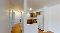 This single-floor unit offers a well-organized layout that balances practicality with comfortable living. Warm hardwood flooring runs throughout the main living areas, hallways, and bedrooms, lending a cohesive and inviting aesthetic to the space. The kitchen is efficiently designed with rich wood cabinetry both above and below the dark granite-style countertops, and is equipped with a white electric range, a stainless steel sink, and a full-size refrigerator. A half-wall pass-through opens the kitchen toward the adjacent living area, enhancing the sense of openness. The living area is generously sized and bathed in natural light through large windows fitted with vertical blinds, which also frame views of surrounding greenery. Baseboard heating and a wall-mounted air conditioning unit ensure year-round comfort. The bedroom features the same hardwood flooring and a bright window with outdoor views. The bathroom includes a wood-toned vanity with a white countertop, a wall mirror with overhead lighting, tile flooring, and a tiled tub-shower combination. A hallway connects the rooms efficiently, with additional closet space noted adjacent to the bathroom. The overall ambiance is clean, functional, and move-in ready.