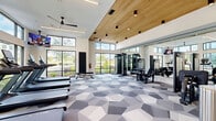 Located at Sawnee Village in Cumming, GA, this fitness center is a spacious, single-floor exercise facility thoughtfully designed to support a full range of workout routines. The architectural design is immediately striking, with soaring ceilings accented by warm wood slat paneling in key zones, creating a refined contrast against crisp white walls. Floor-to-ceiling windows line the perimeter, bathing the entire space in natural light while offering views of the surrounding outdoor greenery and community. Ceiling fans are strategically placed throughout to ensure comfort during intense workouts. The geometric-patterned carpet tile flooring in shades of gray and white adds a contemporary, dynamic aesthetic that flows throughout the room. Equipment is diverse and well-organized, featuring treadmills, stair climbers, stationary cycling bikes with digital screens, ellipticals, and a full cable machine system. A dedicated free weights area includes a comprehensive dumbbell rack and additional strength training benches. Full-length mirrors in the strength training zone enhance spatial perception and allow users to monitor their form. Stability balls and other functional fitness accessories are also present. The facility is further enhanced by wall-mounted TVs and modern pendant lighting, rounding out a premium fitness environment.