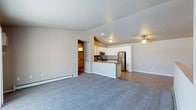 This single-floor space showcases an open-concept design that seamlessly connects the living area to the kitchen, creating a functional and cohesive layout. The expansive living area is finished with plush gray carpeting, providing a comfortable and inviting atmosphere, while recessed lighting illuminates the space evenly.

The kitchen is well-appointed with white cabinetry, granite-look countertops, and a full suite of stainless steel appliances, including a refrigerator, microwave, and range. A peninsula with a breakfast bar serves as both a practical workspace and a casual dining option, effectively separating the kitchen from the living area. The kitchen transitions to wood-look flooring, adding a warm contrast to the surrounding spaces.

A baseboard heating system runs along the living area wall, ensuring efficient climate control. A ceiling fan is visible in the adjacent area, adding to the comfort of the space. An open doorway leads to a warmly lit room, suggesting additional living or sleeping quarters. The neutral color palette throughout creates a clean, versatile backdrop suitable for a variety of interior styles.