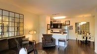 This single-floor space showcases a thoughtfully designed open-concept layout that integrates the living area and kitchen into one cohesive environment. Rich, dark hardwood flooring extends throughout, grounding the space with a sense of warmth and sophistication. The living area is generously appointed, offering room for comfortable seating arrangements, and is enhanced by warm ambient lighting from stylish floor lamps that create an inviting atmosphere. A peninsula-style kitchen counter serves as a natural divider between the living and cooking spaces, providing both a functional workspace and a casual dining or gathering spot. The kitchen features dark upper cabinetry contrasted against light countertops, stainless steel appliances, and pendant lighting that adds a modern touch overhead. The clean white walls throughout serve as a neutral backdrop, allowing the décor and finishes to take center stage. Decorative wall art and framed pieces add personality and visual interest to the space. The layout maximizes the sense of openness while maintaining distinct functional zones, making the space feel both practical and aesthetically refined. Overall, the space exudes a modern, urban ambiance with a cozy residential character.