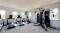This single-level fitness center offers a comprehensive workout environment designed to support a wide range of exercise routines. The space is finished with light gray wood-look flooring throughout, which complements the clean white walls and contributes to a bright, modern aesthetic. Recessed lighting and natural light from numerous windows along multiple walls keep the interior well-illuminated and open.

The cardio zone features multiple Matrix treadmills arranged in a row, an elliptical trainer, a rowing machine, and a stationary spin bike. A wall-mounted flat-screen TV is positioned for visibility during cardio sessions. Adjacent to the cardio equipment is a strength training area anchored by a PowerCage Axis multi-functional cable machine and an Everlast heavy punching bag, catering to both resistance and combat training.

A dedicated free weights area includes a tiered dumbbell rack stocked with a range of weights, alongside an assortment of exercise balls in various sizes for functional fitness and stretching. The open floor plan provides ample circulation space between equipment zones. Windows on multiple sides of the room frame outdoor views, enhancing the overall sense of openness and ventilation throughout the facility.