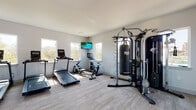 This single-level fitness center offers a comprehensive workout environment designed to support a wide range of exercise routines. The space is finished with light gray wood-look flooring throughout, which complements the clean white walls and contributes to a bright, modern aesthetic. Recessed lighting and natural light from numerous windows along multiple walls keep the interior well-illuminated and open.

The cardio zone features multiple Matrix treadmills arranged in a row, an elliptical trainer, a rowing machine, and a stationary spin bike. A wall-mounted flat-screen TV is positioned for visibility during cardio sessions. Adjacent to the cardio equipment is a strength training area anchored by a PowerCage Axis multi-functional cable machine and an Everlast heavy punching bag, catering to both resistance and combat training.

A dedicated free weights area includes a tiered dumbbell rack stocked with a range of weights, alongside an assortment of exercise balls in various sizes for functional fitness and stretching. The open floor plan provides ample circulation space between equipment zones. Windows on multiple sides of the room frame outdoor views, enhancing the overall sense of openness and ventilation throughout the facility.