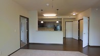 This single-floor residence offers a thoughtfully designed open-concept layout that seamlessly connects the main living areas. The kitchen serves as a central focal point, featuring rich dark wood cabinetry, a tile backsplash, granite countertops, and black appliances including a refrigerator. A raised peninsula provides a natural division between the kitchen and living area while maintaining the open feel of the space. Two pendant lights hang above the peninsula, adding both functional task lighting and a stylish design element, complemented by a flush-mount ceiling fixture nearby. The flooring transitions from rich hardwood-style planks in the kitchen and main living area to plush carpeting in the adjacent room, offering a comfortable contrast underfoot. The layout includes multiple doorways providing access to additional rooms, with a welcoming entry door visible to one side. Neutral wall tones throughout keep the space feeling bright and versatile. The open sightlines and efficient use of space create a cohesive, functional environment well-suited for everyday living and entertaining alike.