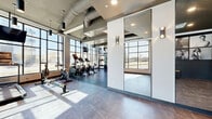 Located at 15 Cooper's Town Road in Winnipeg, this contemporary fitness facility offers a thoughtfully designed, single-level space that combines functionality with modern aesthetics. The facility is organized into distinct zones, each serving a specific purpose within the overall fitness experience.

The main workout area is expansive and sun-drenched, featuring floor-to-ceiling black-framed windows that frame outdoor views and flood the interior with natural light. Exposed ductwork runs along the dark ceiling, lending an industrial character that is balanced by clean white walls and polished tile flooring. Cardio equipment, including treadmills, ellipticals, stationary bikes, and rowing machines, is arranged to take full advantage of the exterior views.

A dedicated strength training zone features rubber flooring, a rack of dumbbells, a cable machine, and additional benching equipment, with pendant and track lighting providing focused illumination throughout.

The entry lobby is equally refined, with large-format marble-effect tile flooring, full-height mirrors framed in sleek black, wall-mounted sconce lighting, and a gallery wall displaying bold black-and-white portrait photography. Restroom facilities are accessible directly from the lobby area, and a glazed door connects the lobby to the main fitness floor, creating a seamless transition between arrival and activity throughout this well-appointed space.