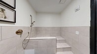 Located at 201 Cayuga Park Lane in Ithaca, NY, this purpose-built pet bathing room is a compact yet thoughtfully designed space dedicated entirely to pet care. The room is finished with large-format, light gray stone-look ceramic tiles that line the lower half of all walls, creating a clean, moisture-resistant surface well-suited for its function. The upper walls are finished in smooth white, lending a bright and airy feel to the space.

At the heart of the room is a built-in bathing tub, elevated on a tiled platform with a set of steps for easy pet access. A brushed gold handheld showerhead and matching wall-mounted valve add a refined, functional touch to the bathing station. The floor is finished in a durable, easy-to-clean light blue surface that contrasts softly with the neutral tile palette.

Framed black-and-white dog illustrations are displayed along the upper wall, adding a welcoming and thematic charm to the otherwise utilitarian space. A wall-mounted electrical outlet is conveniently positioned for grooming tools. The room is accessed through a bold matte black door with a sidelight panel, which introduces a modern, contrasting design element. Warm wood-look flooring is visible just beyond the doorway, hinting at the broader facility this room is part of.