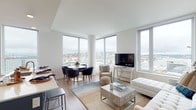 This single-floor high-rise residence is defined by its expansive floor-to-ceiling and corner windows, which frame sweeping panoramic views of the surrounding cityscape and waterfront. The open-concept living and dining area is bathed in natural light, with light hardwood flooring, a neutral color palette, and a layout that comfortably accommodates both lounging and dining. A round dining table with navy upholstered chairs anchors one corner, while the living area features a plush seating arrangement centered around a coffee table and flat-screen TV.

The kitchen is a standout feature, designed with dual-tone navy and white cabinetry, white quartz countertops, a stainless steel gas range, integrated appliances, and a generous island with a double sink — ideal for both everyday cooking and entertaining. A large window in the kitchen extends the city views throughout.

The two bedrooms offer a calm retreat, each with plush carpeting, ample closet space, and windows overlooking the city. One bedroom includes an ensuite bathroom with a glass-enclosed walk-in shower, a backlit vanity mirror, and a white double-drawer vanity. A second full bathroom features a tub-shower combination, completing the well-appointed layout.