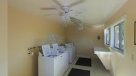 This single-floor shared laundry facility is designed with practicality and functionality in mind. The room features a row of top-loading washing machines lined along one wall, providing ample capacity to accommodate multiple users simultaneously. Informational notices are posted on the walls, ensuring users have easy access to operating instructions and guidelines.

The space is kept comfortable by two ceiling fans with integrated light fixtures, which promote air circulation throughout the room. Natural light filters in through a window, brightening the interior and complementing the warm, neutral-toned walls that give the room a clean and welcoming atmosphere.

A long, built-in counter runs along one side of the room, offering generous surface area for folding laundry or setting down personal items. Dark floor mats are strategically placed to provide traction and protect the tiled flooring in high-traffic areas. The overall layout is thoughtfully organized to maximize usability, with clear pathways between machines and ample room to move comfortably. This laundry facility is a well-maintained, fully functional space that serves the practical needs of its users efficiently.