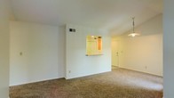 This single-floor residence offers a clean, functional layout designed for comfortable everyday living. The main living area is finished with wall-to-wall carpeting, lending a warm and quiet atmosphere to the space. Crisp white walls throughout enhance the sense of openness and provide a neutral backdrop adaptable to a variety of interior styles. A notable architectural feature is the pass-through window connecting the kitchen to the living area, allowing for easy interaction between spaces while maintaining a sense of defined zones. The kitchen itself is warmly lit, with an inviting glow visible through the pass-through opening. A stylish pendant light hangs in the adjoining area near the entryway, adding a decorative touch and providing focused illumination. HVAC vents are visible along the walls, ensuring year-round climate comfort. The entryway features a solid door and benefits from its own overhead lighting, creating a welcoming transition into the home. Overall, the space presents a practical and well-maintained interior, ideal for those seeking a straightforward, move-in-ready layout with a cohesive and bright aesthetic throughout.