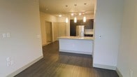 This single-floor residence features an open-concept design that seamlessly connects the living and kitchen areas. The kitchen serves as a focal point, anchored by a central island with a light stone countertop and an integrated sink, offering both workspace and a casual gathering spot. Pendant lighting in a cylindrical style hangs above the island, providing targeted illumination and a modern aesthetic touch. Additional track lighting on the ceiling further brightens the space.

The kitchen is equipped with dark-toned cabinetry that extends to the upper and lower sections, complemented by a stainless steel refrigerator and a tile backsplash that adds a subtle textural element. The open layout allows the kitchen to flow naturally into the surrounding living space.

Throughout the unit, dark hardwood-style flooring creates a rich, cohesive base that contrasts with the light, neutral-toned walls. The clean white baseboards and smooth wall surfaces contribute to a contemporary, well-maintained atmosphere. Multiple doorways visible from the main area suggest additional rooms beyond, while the overall layout offers a practical and modern living environment.