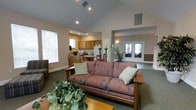 This single-story space features an open-concept design that seamlessly connects the living area, kitchen, and an adjoining room. The living room is anchored by a patterned sofa with accent pillows and a Craftsman-style wooden side table, complemented by lush indoor plants that add a natural, welcoming touch. Neutral-toned carpet covers the floor throughout the main living space.

A prominent architectural feature is the vaulted ceiling over the living area, fitted with recessed lighting that provides consistent illumination. Large windows along one wall allow natural light to fill the space, enhancing its open feel.

The kitchen, visible from the living area, includes warm wood cabinetry, a white refrigerator, countertop appliances, and a breakfast bar with seating. The kitchen area transitions naturally into a small dining space.

Toward the rear of the space, a set of French doors with glass panels opens into a separate room, which appears to be a sunlit enclosure or additional gathering area. Decorative candle stands and potted greenery provide tasteful accents throughout, contributing to the overall warm and comfortable ambiance of the space.