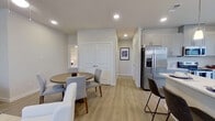 This single-story residence offers a thoughtfully designed layout across two bedrooms and two bathrooms. The open-concept living, dining, and kitchen area serves as the heart of the home, featuring light hardwood-style flooring, recessed lighting, and neutral tones throughout. The kitchen is well-appointed with gray shaker cabinetry, white countertops, stainless steel appliances including a French door refrigerator with ice maker, and a peninsula with bar seating illuminated by pendant lighting. A sliding glass door in the kitchen area provides direct access to a private outdoor patio, bringing in abundant natural light. The dining area accommodates a round table and is positioned near a double-door closet for additional storage. The living room features a large window that fills the space with natural light. The primary bedroom includes a walk-in closet with built-in shelving and rods. The en-suite bathroom features a glass-enclosed walk-in shower, gray vanity cabinetry, and white countertops. The second bedroom offers carpeted flooring and a bright window. The consistent neutral palette and modern finishes create a clean, cohesive aesthetic throughout the residence.