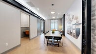 This professionally designed business center suite offers a thoughtfully curated single-floor layout ideal for meetings, collaboration, and focused work. The central conference area is anchored by a large rectangular table surrounded by sleek black leather chairs, illuminated by striking geometric pendant lights that hang from a bright, recessed-lit ceiling. Underfoot, light-toned herringbone hardwood flooring extends throughout, adding warmth and visual interest to the space.

A defining feature is the large-scale vintage-inspired mural wall, which displays a Guglielmo Marconi quote and illustration, lending the space a creative and intellectual character. Adjacent to it, an exposed stacked-stone accent wall adds rich textural contrast alongside dark industrial steel columns.

Floor-to-ceiling windows line one side of the suite, flooding the space with natural light and offering views of the surrounding streetscape and greenery. A glass-partitioned lounge area provides a more relaxed setting, furnished with vibrant amber leather armchairs and a round side table — perfect for informal conversations. A separate enclosed workstation with its own sliding door offers a private, focused workspace. The combination of modern fixtures, industrial accents, and thoughtful spatial separation makes this suite a highly functional and visually engaging professional environment.