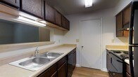 This single-floor residential unit offers a straightforward and functional layout designed for comfortable everyday living. The main living area features a combination of dark hardwood flooring and carpeting, creating distinct zones within the open space. Built-in shelving lines one wall, providing convenient display or storage options, while a window along the far wall allows natural light to brighten the room. The neutral wall tones and textured ceiling contribute to a clean, understated aesthetic throughout.

A hallway connects the living area to the rest of the unit, leading toward additional rooms at the rear. The kitchen is efficiently designed, featuring rich dark wood cabinetry that extends to the upper walls for generous storage capacity. Light-toned countertops provide ample workspace, complemented by a double stainless steel sink. Under-cabinet lighting adds functional illumination to the prep area, and light wood-style flooring ties the space together. A door off the kitchen offers access to an adjacent area, adding to the overall connectivity of the layout.

Overall, this unit presents a practical and well-organized living environment with consistent finishes and a layout that makes efficient use of its single-floor footprint.