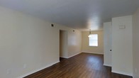 This single-story space features an open layout connecting the living and dining areas, creating a cohesive and functional flow throughout. Dark hardwood flooring runs continuously across the space, providing a rich contrast against the neutral, light beige walls and white baseboards. The textured ceiling adds a subtle architectural detail overhead.

The dining area is defined by a classic pendant light fixture that hangs centrally, drawing the eye and providing focused illumination. A window positioned in this area allows natural light to filter into the space, brightening the interior. HVAC vents are integrated into both the ceiling and walls, ensuring efficient climate control throughout.

An open passageway connects the two areas seamlessly, maintaining an airy and unobstructed feel. The overall layout is straightforward and versatile, lending itself well to a variety of furniture arrangements. The combination of dark flooring, warm neutral tones, and clean lines gives the space a simple yet inviting ambiance suitable for everyday living.