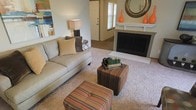 This single-floor residence centers around a welcoming living area designed with comfort and style in mind. The living room serves as the heart of the space, anchored by a classic fireplace with a clean white mantel, decorated with striking orange vases and a decorative sunburst mirror that adds a contemporary focal point. Plush wall-to-wall carpeting in a warm neutral tone grounds the room, complementing the light gray sofa adorned with coordinating throw pillows. A pair of striped ottomans in earth tones adds both functionality and visual interest to the seating arrangement. A tasteful table lamp provides warm ambient lighting, while colorful abstract wall art introduces a vibrant accent to the otherwise neutral palette. Natural light filters into the space through windows dressed with blinds, visible toward the rear of the room near what appears to be an entryway door. The open layout allows for easy flow between areas, and the combination of soft textiles, warm accent colors, and thoughtful décor creates a cohesive and inviting atmosphere throughout.