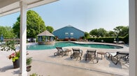 Located at 238–244 Kyser Blvd in Madison, Alabama, this outdoor amenity complex centers around a generously sized freeform swimming pool with a distinctive curved design. The pool's clear, blue-green water is complemented by a striking stone-clad gazebo situated directly within the pool area, providing a shaded gathering spot accessible from the pool deck. Surrounding the pool is a broad, smooth concrete sun deck furnished with multiple lounge chairs and side tables, offering ample space for relaxation and sunbathing. A covered patio area adjoins the main building, which features classic brick construction with teal-painted doors and a pitched shingle roof. This shaded corridor provides additional sheltered outdoor space with open views toward the pool and fenced grounds. Mature trees and well-maintained landscaping border the entire area, creating a private, resort-like atmosphere. Additional seating areas with patio tables and umbrellas are positioned poolside, ideal for outdoor dining or social gatherings. Safety signage, including pool rules and a no-lifeguard notice, is clearly posted on the building exterior. The overall design blends functional amenities with attractive landscaping, delivering a welcoming and well-appointed outdoor recreational environment.