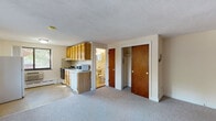 Located at 15 Chester St in Cambridge, MA, this single-floor unit offers a practical and straightforward layout designed for comfortable everyday living. The main living area is generously sized, finished with neutral wall-to-wall carpeting that runs throughout, and receives natural light through a large picture window fitted with baseboard heating below. A full-size refrigerator anchors one corner of this open space.

The kitchen is efficiently arranged along one wall, featuring warm honey-toned oak cabinetry both above and below the countertops, a white tile backsplash, a stainless steel sink, an electric range with oven, a dishwasher, and a microwave. A window above the counter brings in additional natural light, creating a bright and functional cooking environment.

The bedroom is a well-proportioned room distinguished by a bold dark wood-paneled accent wall, soft carpeting, and two closets with wooden doors, offering ample storage. A textured ceiling is present throughout the unit.

The bathroom is clean and simply appointed, featuring full white subway tile surrounding a soaking bathtub with a window above, providing ventilation and light. The layout is compact and efficient. Overall, the unit presents a functional, no-frills living space in a well-situated Cambridge location.