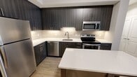 This well-designed three-bedroom, two-bathroom residence in Alexandria, Virginia offers a thoughtfully laid-out single-floor plan with modern finishes throughout. The open-concept kitchen and living area serves as the heart of the home, showcasing dark wood-grain cabinetry that extends floor to ceiling, crisp white quartz countertops, a neutral tile backsplash, and a full suite of stainless steel appliances including a range, microwave, and dishwasher. A center island provides additional prep and dining space, while wood-look flooring ties the area together seamlessly. The three bedrooms feature plush carpeting and neutral tones, with natural light filtering in through window blinds. The generously sized primary bedroom includes access to a dedicated walk-in closet outfitted with wire shelving and hanging rods for ample storage. The bathrooms are finished with clean, contemporary details, highlighted by a frameless sliding glass shower enclosure set against large-format white tile. A hallway connects the living spaces efficiently, and an additional closet provides further storage. The overall ambiance is clean, modern, and move-in ready, reflecting a cohesive design aesthetic suited for comfortable everyday living in the heart of Alexandria.