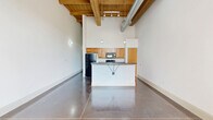 Located at 2100 Main Street in Porterdale, GA, this single-floor loft-style unit showcases an industrial-chic aesthetic with a distinctive character. Soaring ceilings with exposed wooden beams and visible ductwork set a dramatic tone throughout the space, while polished concrete floors provide a sleek, durable foundation underfoot.

The open-concept kitchen is a central feature, offering warm honey-toned cabinetry, dark granite-style countertops, and a full suite of black appliances including a refrigerator, electric range, and built-in microwave. A peninsula with a breakfast bar provides additional prep space and casual seating. Industrial-style pendant lighting illuminates the kitchen area with character.

A long hallway connects the living and sleeping areas, leading to a bedroom that showcases a stunning exposed brick wall with a large arched window, flooding the room with natural light. A ceiling fan adds comfort to this architecturally rich space.

The bathroom features tile flooring, a tub-shower combination with full tile surround, and a double-sink vanity with framed mirrors. Two closets offer storage, and an in-unit laundry closet with a full-size washer and dryer adds everyday convenience. This space blends historic industrial charm with modern functionality.