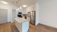 Located at 801 Watkins View Dr in Midlothian, Virginia, this single-floor residence offers a well-designed layout with modern finishes throughout. The kitchen serves as a centerpiece of the home, showcasing crisp white shaker-style cabinetry, granite countertops with subtle speckled detailing, and a full suite of stainless steel appliances including a refrigerator, dishwasher, microwave, and range. A kitchen island with a matching granite surface provides additional prep space and is accented by pendant lighting overhead, while warm wood-look flooring ties the space together. The open flow from the kitchen into the living area creates an inviting, functional layout. The living area features plush neutral carpeting, offering a comfortable contrast to the hard flooring in the kitchen and hallways. A ceiling fan with built-in lighting and large windows with exterior views enhance the room's brightness and comfort. The bathroom is well-appointed with white cabinetry, granite countertops, a double vanity with large mirrors, and a full soaking tub. Wood-look flooring extends through the bathroom, and the space is connected to a carpeted bedroom area beyond, completing this thoughtfully finished single-level home.