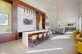This single-floor communal space presents a refined, upscale design characterized by dramatic soaring ceilings that create an expansive, airy atmosphere throughout. The central focal point is a sleek bar counter with a crisp white surface and rich wood-toned base, accommodating several cushioned bar stools and backed by a striking marble-clad wall with dark pendant lighting cascading at varying heights. A mounted flat-screen television is integrated into this feature wall, enhancing both form and function.

Adjacent to the bar, a comfortable lounge area is furnished with neutral-toned armchairs and a glass-top coffee table, anchored by a warm area rug. Wall paneling and high mounted artwork contribute a polished, gallery-like quality. Further into the space, a dining area with high-top tables and colorful upholstered chairs sits beneath a sculptural sputnik chandelier.

Along one wall, floor-to-ceiling windows with decorative drapery introduce abundant natural light and outdoor views, complemented by additional seating with vibrant mustard-yellow accent chairs. Light wood-tone flooring runs consistently throughout, tying the various zones together into a cohesive, welcoming environment designed for socializing and relaxation.