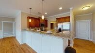 Located at 7205 Wrightsville Ave in Wilmington, NC, this single-floor residence offers a well-designed open-concept layout that seamlessly connects the living and kitchen areas. The kitchen is a standout feature, showcasing rich medium-toned wood cabinetry, granite countertops, a large peninsula with seating, stainless steel appliances including a refrigerator, range, and microwave, and warm hardwood flooring. Pendant lighting adds a refined touch above the peninsula, while crown molding throughout elevates the overall finish.

The expansive living area is bright and airy, benefiting from large windows and sliding glass doors that invite abundant natural light and provide views of the surrounding area. Wainscoting panels along the lower walls and a ceiling fan add classic architectural character to this generous open space.

The bedrooms are carpeted for comfort, each offering ceiling fans and access to outdoor views via sliding glass doors. The bathrooms are clean and functional, featuring hardwood-style flooring, a tub-shower combination, and brushed nickel fixtures. The overall ambiance is warm and inviting, with neutral tones, consistent finishes, and thoughtful detailing throughout.